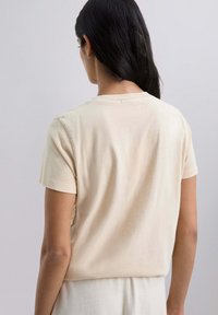Camiseta de manga corta beige en suave algodón con un cuello redondo, que presenta un corte holgado y una textura suave, mostrada desde la parte de atrás.