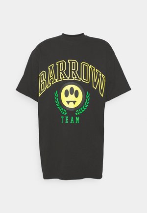 Musta ylisuuri T-paita, jossa on keltainen "BARROW TEAM" -teksti, vihreä hymyilevä logo ja neonvihreä hihna. Yhdistettyjen löysien mustien housujen ja mustien kenkien kanssa.