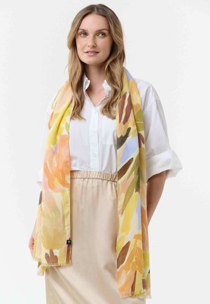 Woman with light brown hair wearing a white shirt and beige skirt, draped in a large floral scarf with yellow, orange, and green tones.