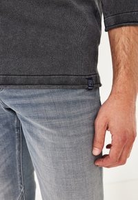 Dunkelgrauer Strickpullover mit einem kleinen Markenpatch, kombiniert mit hellblauen Jeans, die eine lockere Passform und subtile Texturen zeigen.