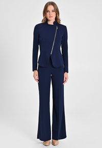 Ensemble deux pièces bleu marine composé d'une veste ajustée avec une fermeture éclair dorée diagonale et un pantalon à jambes larges. Tissu lisse avec un design structuré.