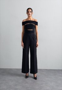 Top negru cu umeri descoperiți, cu decupaje, asortat cu pantaloni largi negri cu talie înaltă și tocuri negre, pe un fundal neutru.