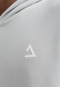 Grå hoodie med en mjuk textur, med en vit logotyp i form av en triangel och texten "JACK & JONES CORE" längst ner.