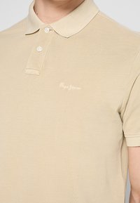 Bežová polokošeľa s krátkym rukávom, s dvoma bielymi gombíkmi a diskrétnym výšivkou "Pepe Jeans" na hrudi, nosená osobou.