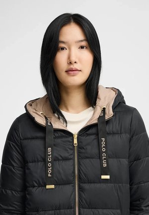 Vrouw met steil zwart haar, gekleed in een zwart doorgestikt jack met een beige capuchon en zwarte trekkoorden met het label "Polo Club".