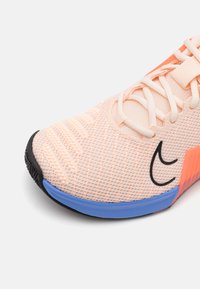 Zapatilla atlética ligera con una parte superior de malla transpirable en durazno pálido con acentos en negro y una suela azul y naranja. Superficie texturizada.