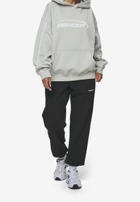 Sudadera con capucha gris claro con un bolsillo delantero y el logo blanco "PEGADOR". Combinada con pantalones deportivos negros que presentan un pequeño logo. Se ven unas zapatillas blancas.