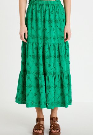 Zielona spódnica midi na falbanach z kwiatowym haftem, ażurowymi detalami i elastycznym paskiem, noszona z brązowymi sandałami na paski na białym tle.
