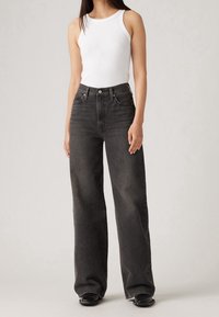 Černé džíny s vysokým pasem a širokými nohavicemi, vyrobené z denimu, s předními kapsami a zapínáním na knoflíky, v kombinaci s upnutým bílým tílkem.