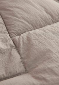 Nahaufnahme einer hellbeigen, gesteppten Bettwäsche mit einer zerknitterten Textur, auffälligen Nähten und weichen, gepufften Abschnitten, die eine Komfortschicht bilden.