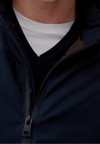 Marineblå lynlåsjakke med høj krave, der har en kontrasterende inderside og en sort lynlås. Båret over en hvid crew neck skjorte.