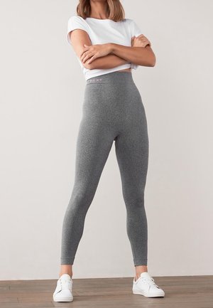 Leggings gris taille haute avec une texture côtelée, ornés d'une bande logo « NEXT » rose en haut, associés à des baskets blanches.
