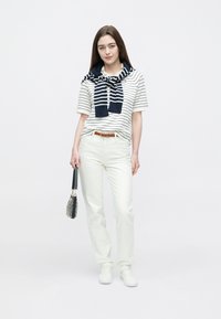 Fehér és tengerészkék csíkos rövid ujjú galléros póló, tengerészkék pulóverrel a nyak körül; fehér nadrággal és sportcipővel párosítva.