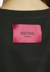 Schwarzes Stoffkleidungsstück mit einem pinken Satinlabel. Der Text auf dem Label lautet "NICOWA MADE IN ITALY". Glatte Textur und minimalistisches Design.