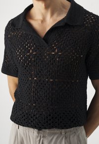 Persona que lleva una camisa de crochet negra de manga corta con cuello, combinada con pantalones cortos de color beige claro, mostrada desde el cuello hasta la cintura.