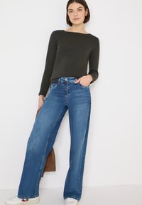 High-waisted, wijdbenige blauwe spijkerbroek met een verweerd effect, gecombineerd met een aansluitende donkergroene trui met lange mouwen. Witte schoenen en een bruin accessoire inbegrepen.