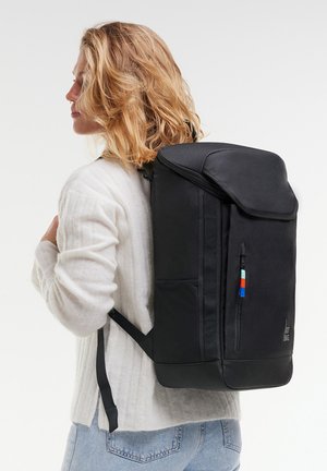 Personne blonde portant un pull blanc et un jean bleu clair, portant un grand sac à dos noir avec une fermeture éclair à l'avant et un onglet de tirage coloré.