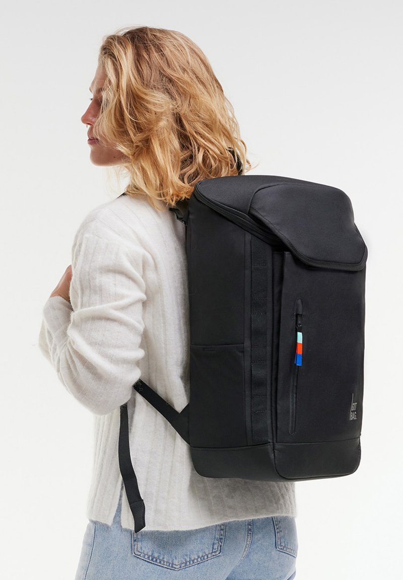 Persona rubia que lleva un suéter blanco y jeans azul claro, con una mochila grande y negra con cremallera frontal y una lengüeta colorida.