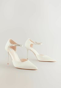 Valkoiset korkeakorkoiset kengät, joissa on kapea kärki, sileä kangas, nilkkaremmi metalliakselilla ja hoikka stiletto-korko.