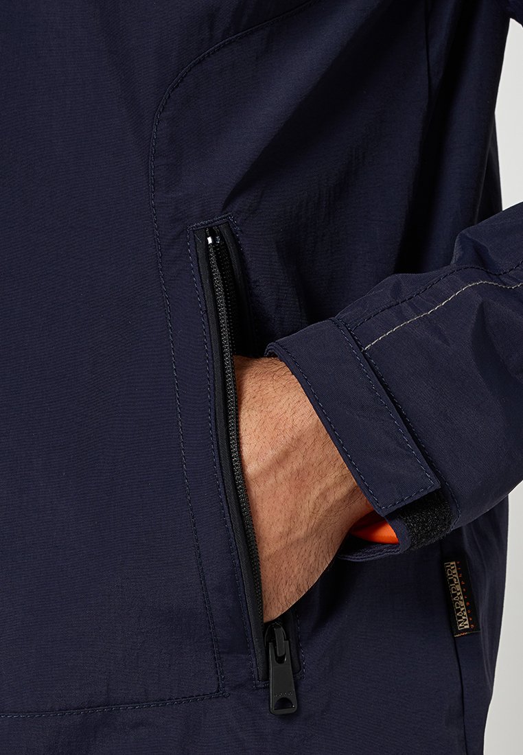 Giacca blu navy con tasca con zip, tessuto testurizzato e cuciture a contrasto. Include un accento arancione all'interno della tasca.