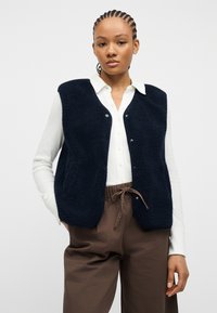 Νεαρή γυναίκα με πλεξούδες, φοράει μπλε φλις γιλέκο πάνω από λευκό πουκάμισο με κουμπιά και καφέ παντελόνι με κορδόνια, στέκεται μπροστά σε απλό φόντο.