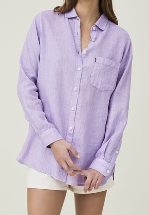 Femme portant une chemise lavande à manches longues avec une poche poitrine et un short blanc, debout les mains doucement jointes devant.