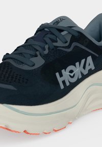 Κοντινό πλάνο ενός μαύρου και γκρι τρέξιμου παπουτσιού HOKA με παχιά λευκή σόλα και πορτοκαλί πέλμα, που δείχνει τα κορδόνια και το λογότυπο της μάρκας στο πλάι.