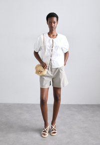 Bluză albă cu mâneci bufante și detaliu cu șnur, asortată cu pantaloni scurți gri deschis și sandale espadrille cu curea bej. Geantă mică țesută.