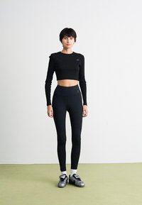 Kvinna med kort mörkt hår som bär en svart långärmad cropped topp, högmidjade svarta leggings, vita strumpor och silverfärgade sneakers, stående på ett grönt golv.
