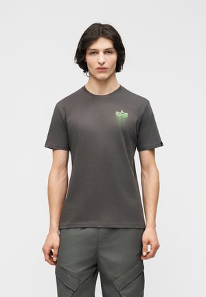 Νέος άνδρας με μεσαίου μήκους σκούρα μαλλιά που φοράει εφαρμοστό σκούρο γκρι T-shirt με ένα μικρό πράσινο φωτεινό αστέρι στο στήθος.