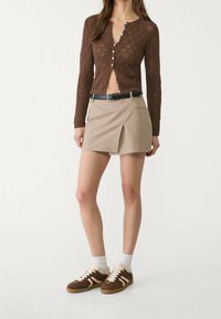 Cardigan in maglia marrone con chiusura a bottone, abbinato a una mini gonna plissettata beige e sneakers in camoscio marrone con dettagli bianchi.