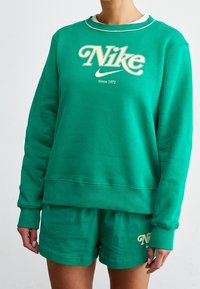 Grön sweatshirt med rundad hals och ribbade ärmmuddar, med en stor "Nike"-logotyp i krämfärg. Kombinerat med matchande gröna shorts.