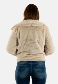 Chaqueta de piel sintética beige con un collar ancho, superficie texturizada y un corte relajado, combinada con vaqueros de mezclilla azules que tienen bolsillos decorativos en la parte trasera.