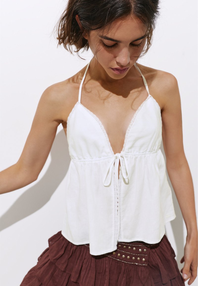 Jeune femme portant un haut blanc dos nu avec un lien sur le devant et une jupe marron à plusieurs couches avec des détails cloutés, regardant vers le bas avec les cheveux attachés.