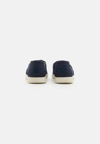 Marineblå lerret slip-on sko med teksturert finish, beige gummisåler og en liten logo på baksiden. Minimalistisk design med lav profil.