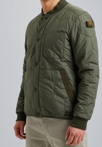 Quilted olivgrüne Jacke mit Knopfleiste, gerippten Bündchen und Seitentaschen. Ausgestattet mit einem strukturierten Stoff und einem akzentuierten Logopatch.