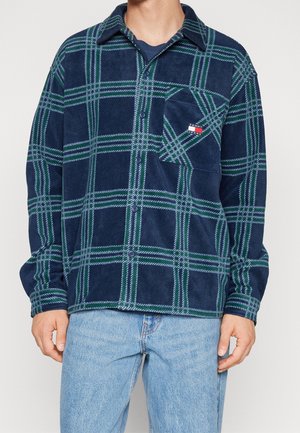 Mężczyzna w niebiesko-zielonej koszuli z flaneli w kratę na guziki z przednią kieszenią, w połączeniu z jasnoniebieskimi jeansami.