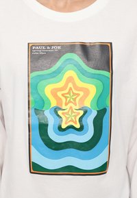 Hvit bomulls T-skjorte med et flerfarget grafisk trykk med lagdelte former i grønt, gult og blått, rammet inn av en svart kant.