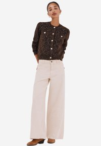 Cardigan marrone a pois con bottoni, indossato sopra pantaloni beige a gamba larga, abbinato a stivaletti a tacco marroni. Design semplice e moderno.