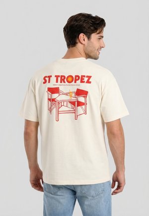 Hombre con camiseta crema con texto rojo "ST TROPEZ" y gráfico de dos sillas y una mesa con una bebida, combinado con vaqueros azul claro.