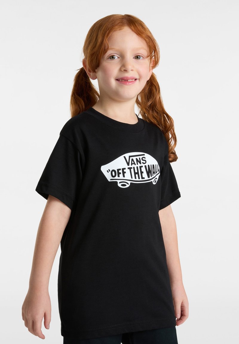 Malá dívka s červenými vlasy, které má v culících, oblečená v černém tričku Vans "Off The Wall", usmívá se, stojí na bílém pozadí.
