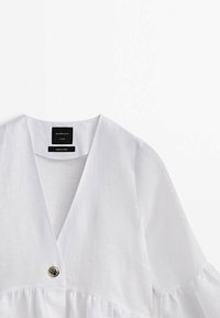 Blouse en lin blanche avec col en V profond, fermeture à un bouton et détail froncé à la taille, présentée sur fond blanc.