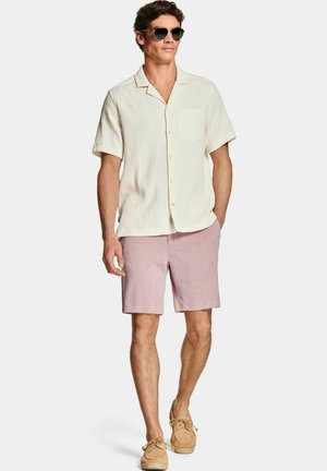Mann mit Sonnenbrille, cremefarbenem kurzärmligem Hemd mit Knöpfen, pinken Shorts und beigefarbenen Schnür-Loafers steht vor einem schlichten Hintergrund.