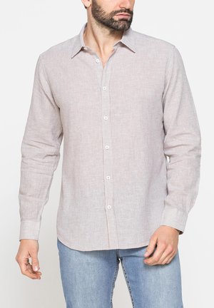 Uomo che indossa una camicia beige chiaro a maniche lunghe con bottoni e jeans azzurri chiari, in piedi davanti a uno sfondo bianco semplice.