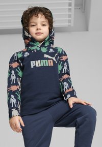 Sudadera con capucha de tela azul oscura, que presenta un patrón multicolor de animales y figuras en las mangas y un logo central de "PUMA" en beige y verde.