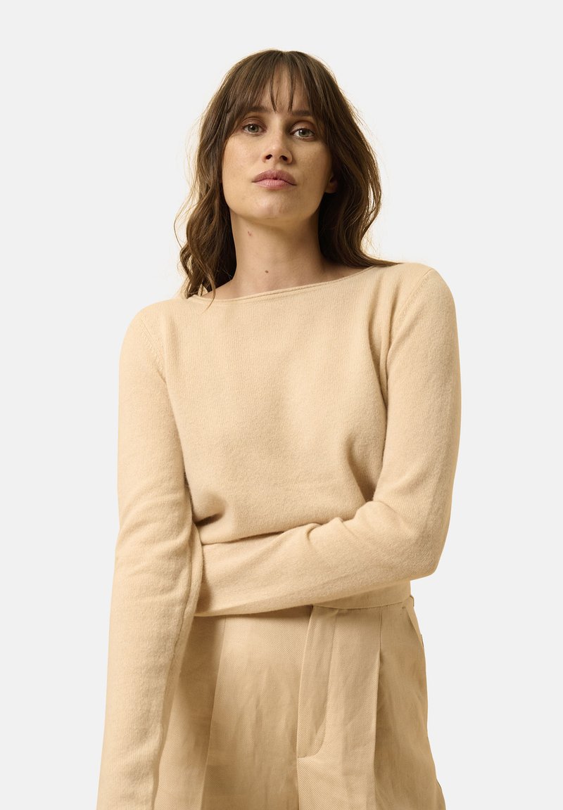 Beiger Langarmpullover aus weichem Material mit einem Schiffsdekolleté und gerippten Details. Das Modell trägt ihn mit beigen hoch taillierten Hosen.