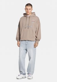 Brązowy hoodie z przednią kieszenią, sznurkami i haftowanym napisem; w połączeniu z jasnoniebieskimi luźnymi dżinsami i białymi trampkami z akcentami.