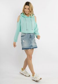 Hoodie cropped di un turchese chiaro con design a spalle scoperte, abbinato a una gonna di jeans strappata e sneakers bianche con accenti gialli.