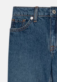 Temno modri jeans iz denima z oranžnim šivanjem, sprednjim žepom in kovinsko zaponko na pasu. Vidna teksturirana tkanina.
