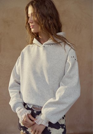 Fiatal nő hosszú, hullámos hajjal világosszürke kapucnis pulóvert visel fém szegecsekkel, fehér fodros ujjvég gumikkal és sötét mintás nadrággal, balra nézve.